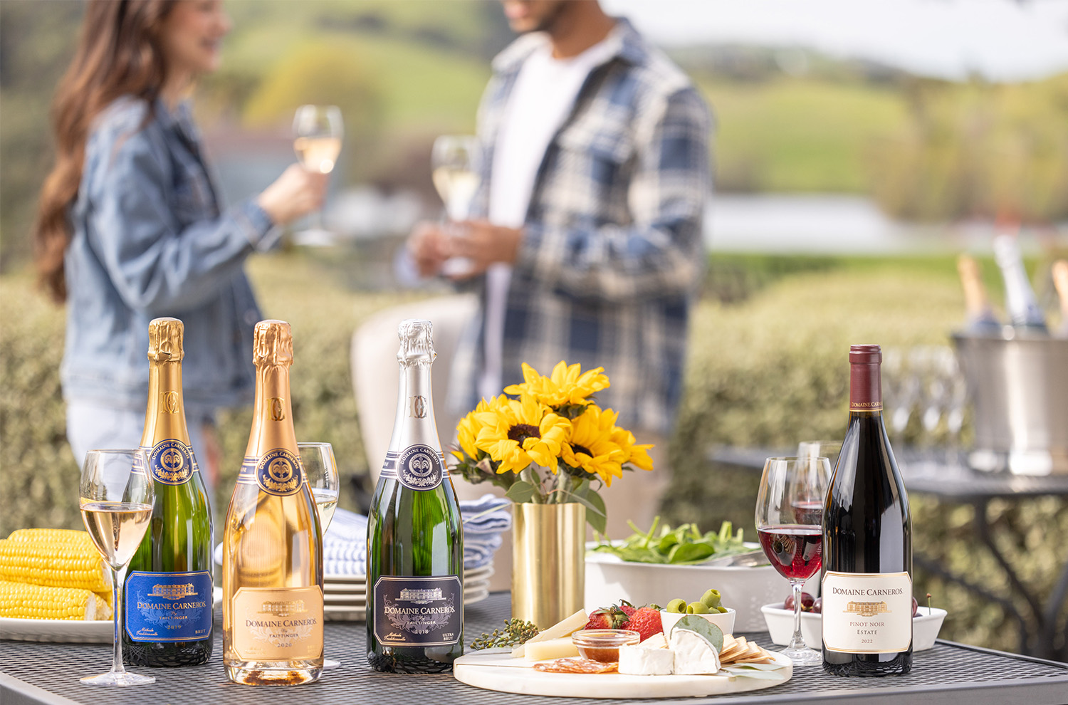 outdoor table set with 4 bottles of wine, vase of sunflowers, cheeseboard, and 2 people blurred in the background holding glasses